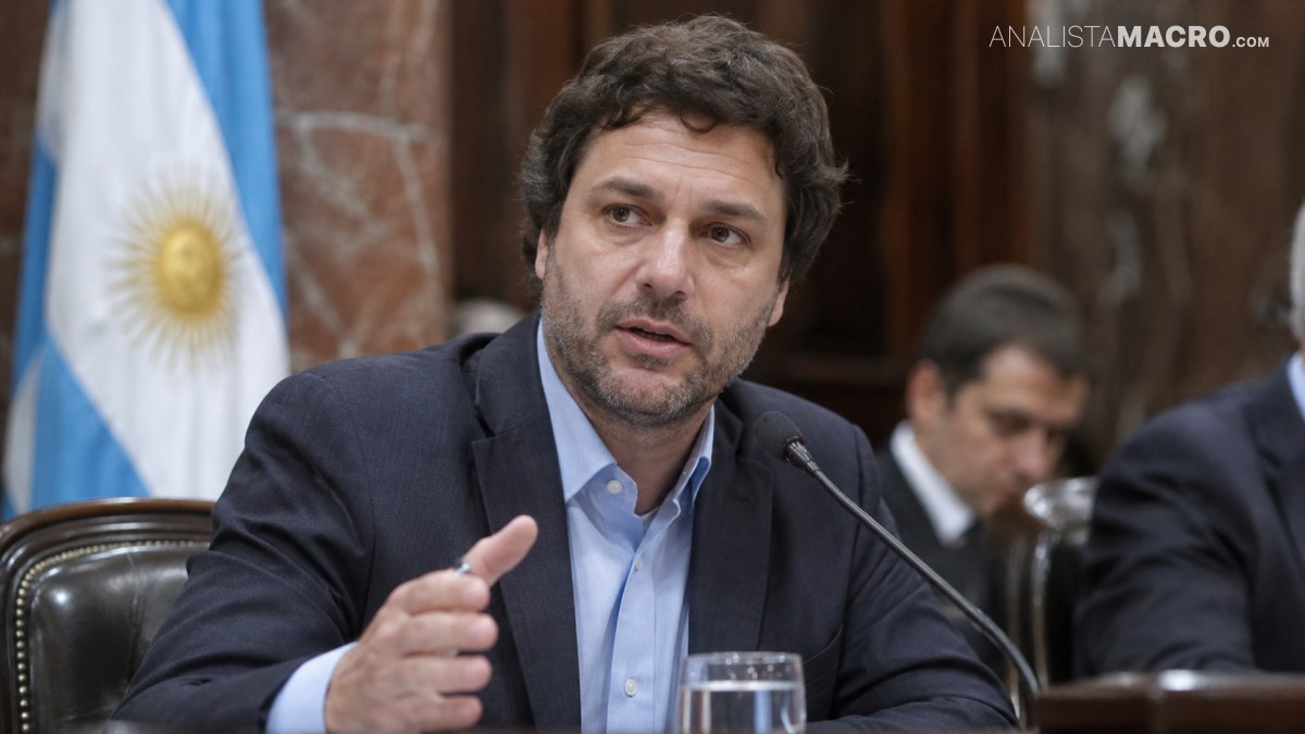 Hombre de cabello oscuro habla ante un micrófono, sentado junto a una mesa con un vaso de agua.