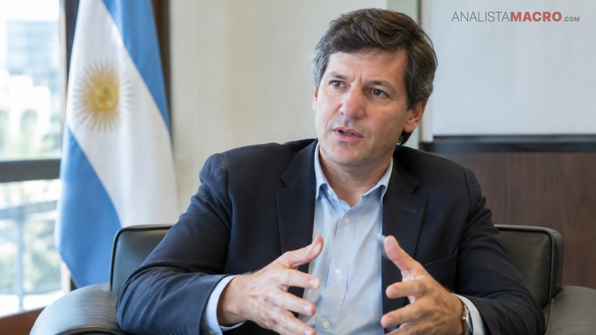 Hombre de traje oscuro y camisa celeste sentado, gesticulando frente a una bandera argentina.