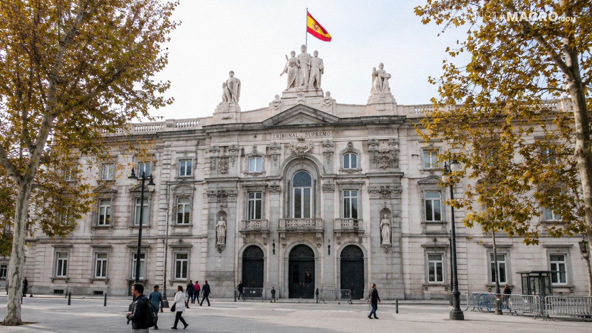 Fachada de un edificio monumental de piedra, con esculturas en la azotea, una bandera española y árboles otoñales a ambos lados.
