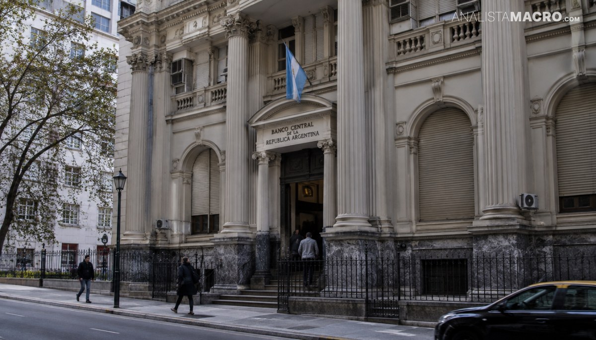Edificio histórico con columnas, entrada central y bandera celeste, rodeado de rejas, árboles, peatones y un automóvil negro.