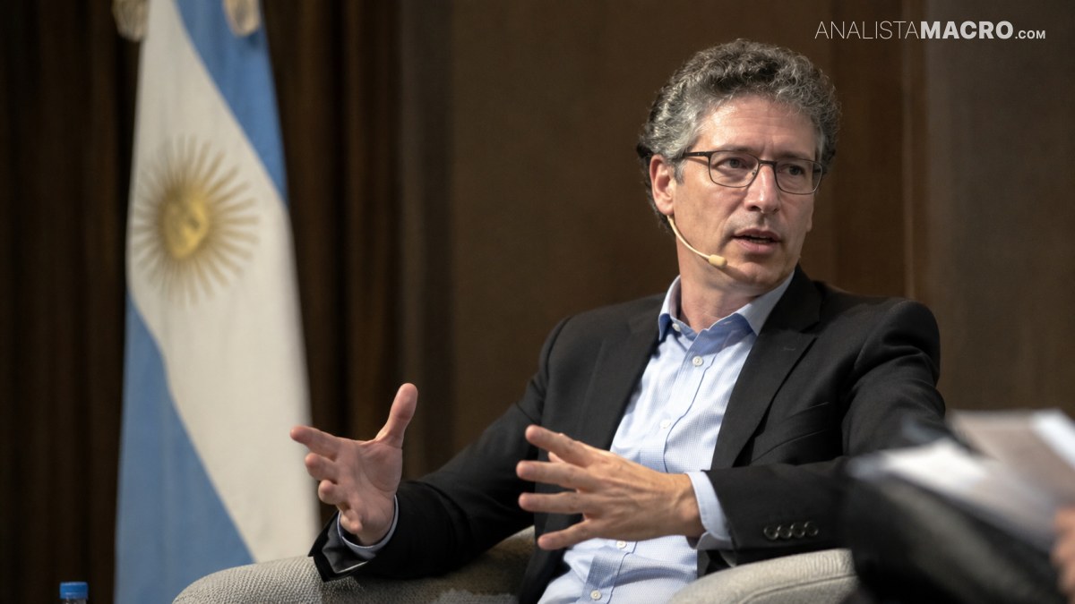 Hombre con gafas y traje negro gesticula sentado en un sillón, con una bandera argentina al fondo.