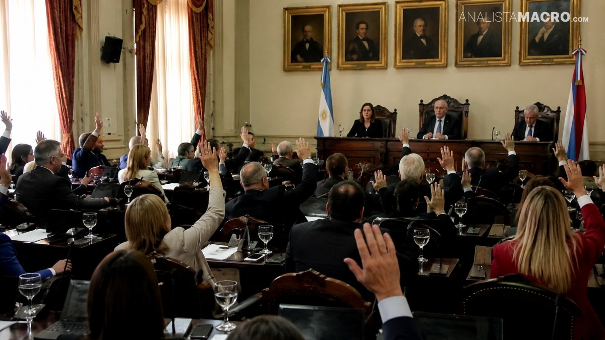 Personas reunidas en una sala formal levantan las manos, mientras varias autoridades están sentadas al frente bajo retratos y banderas.