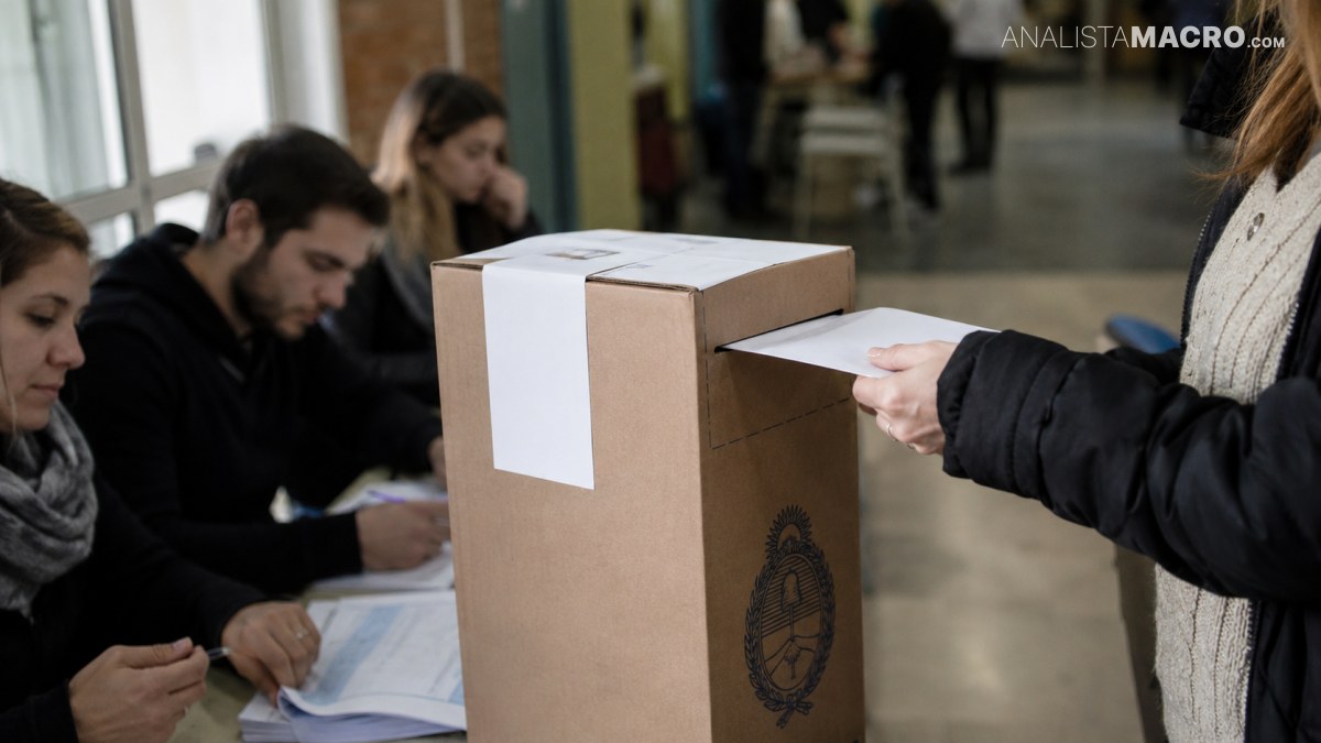 Una persona introduce una papeleta en una urna de cartón mientras varias personas esperan sentadas junto a documentos.