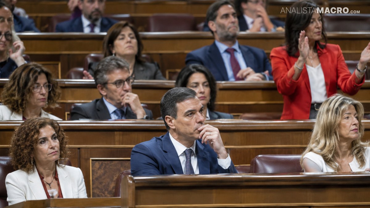 Varias personas están sentadas en escaños de madera de una cámara parlamentaria, mientras algunas aplauden y otras observan.