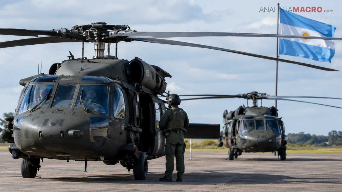 Dos helicópteros militares estacionados en una pista, con una persona uniformada junto al helicóptero delantero y banderas ondeando al fondo.