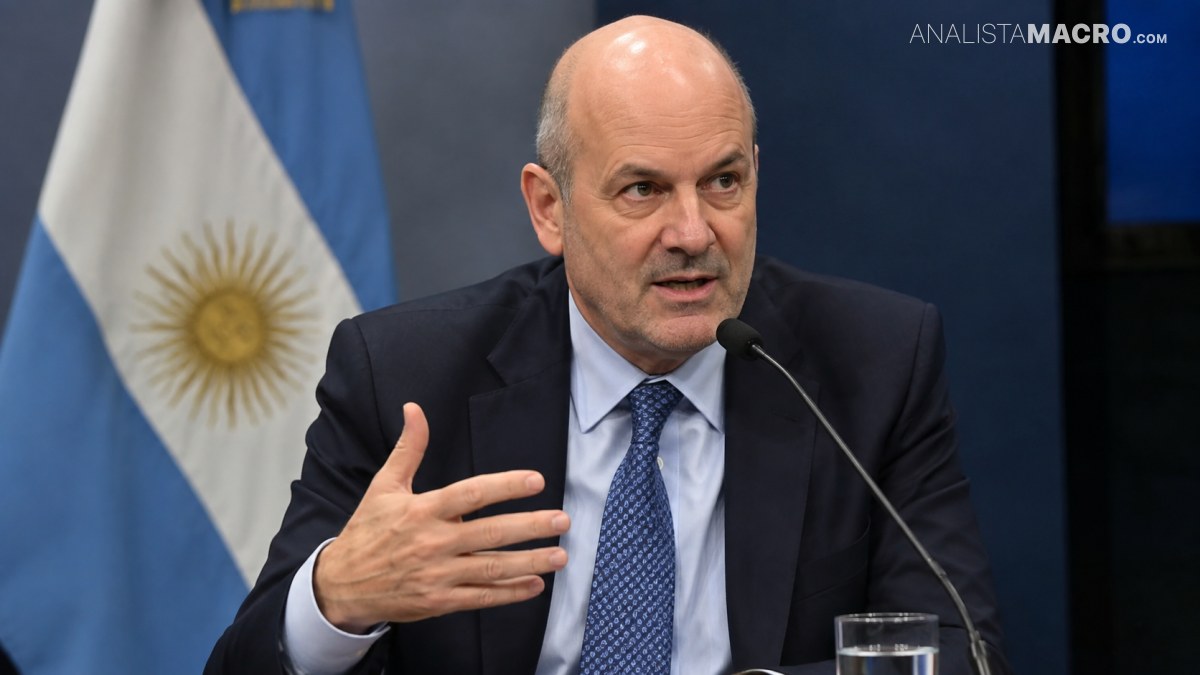 Hombre calvo con traje y corbata habla ante un micrófono, gesticulando con una mano, frente a una bandera argentina.