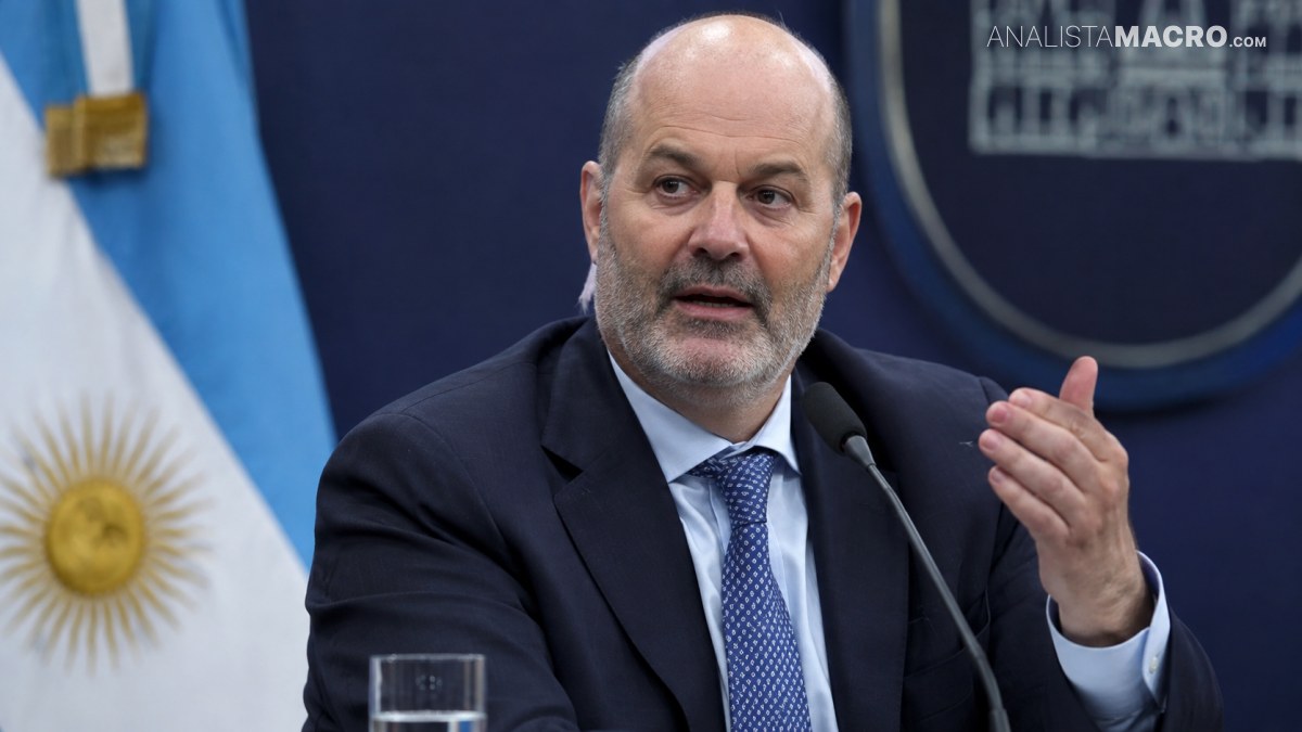 Hombre calvo con barba, traje oscuro y corbata, habla ante un micrófono mientras gesticula frente a una bandera argentina.