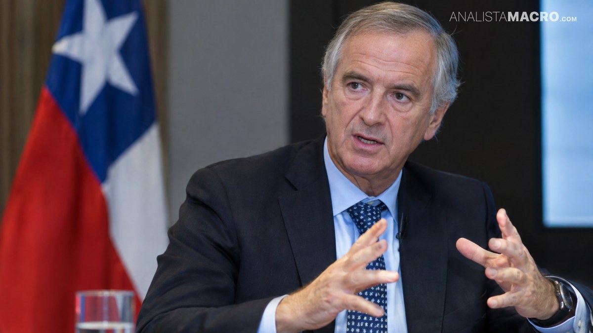 Hombre de cabello gris con traje oscuro, hablando y gesticulando frente a una bandera y una pantalla.