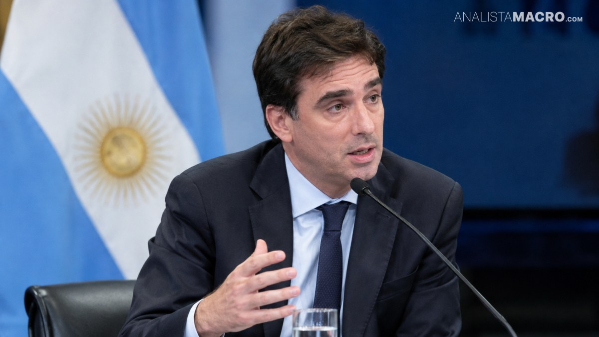 Hombre con traje oscuro habla en un atril, sostiene la mano al frente y hay una copa con agua.