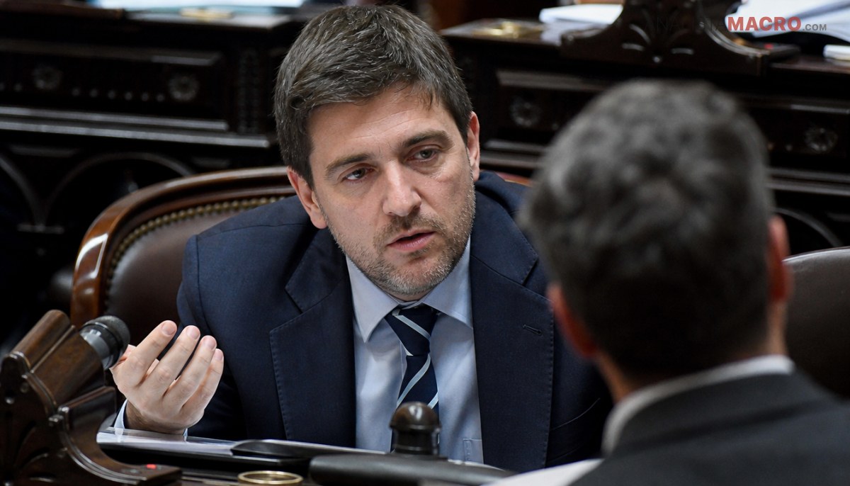 Hombre con traje oscuro hablando y gesticulando en una reunión, con micrófono y papeles al fondo.
