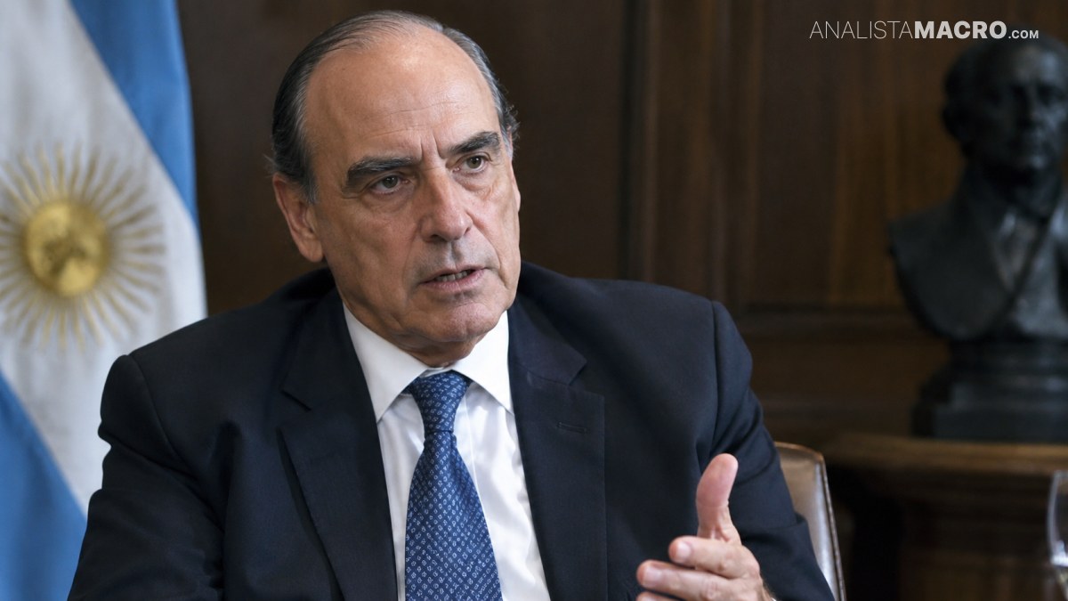Hombre con traje oscuro y corbata azul gesticula mientras está sentado, con una bandera argentina y un busto al fondo.