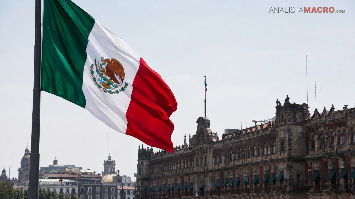 La bandera mexicana ondea frente a un edificio histórico de varios pisos, con un cielo despejado al fondo.