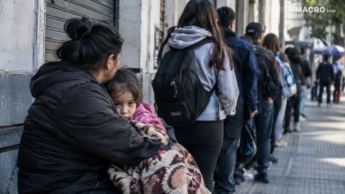 Mujer sentada abraza a una niña pequeña envuelta en una manta, mientras varias personas hacen fila junto a un edificio.
