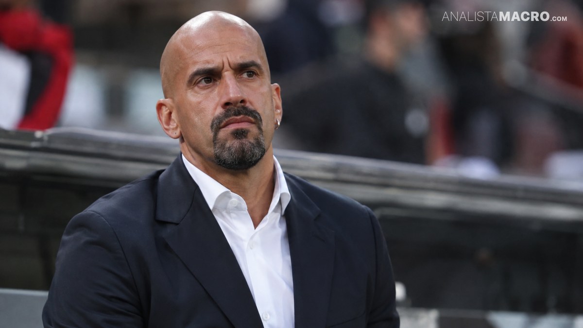 Hombre calvo con barba y traje negro, mirando serio desde un estadio con el fondo desenfocado.