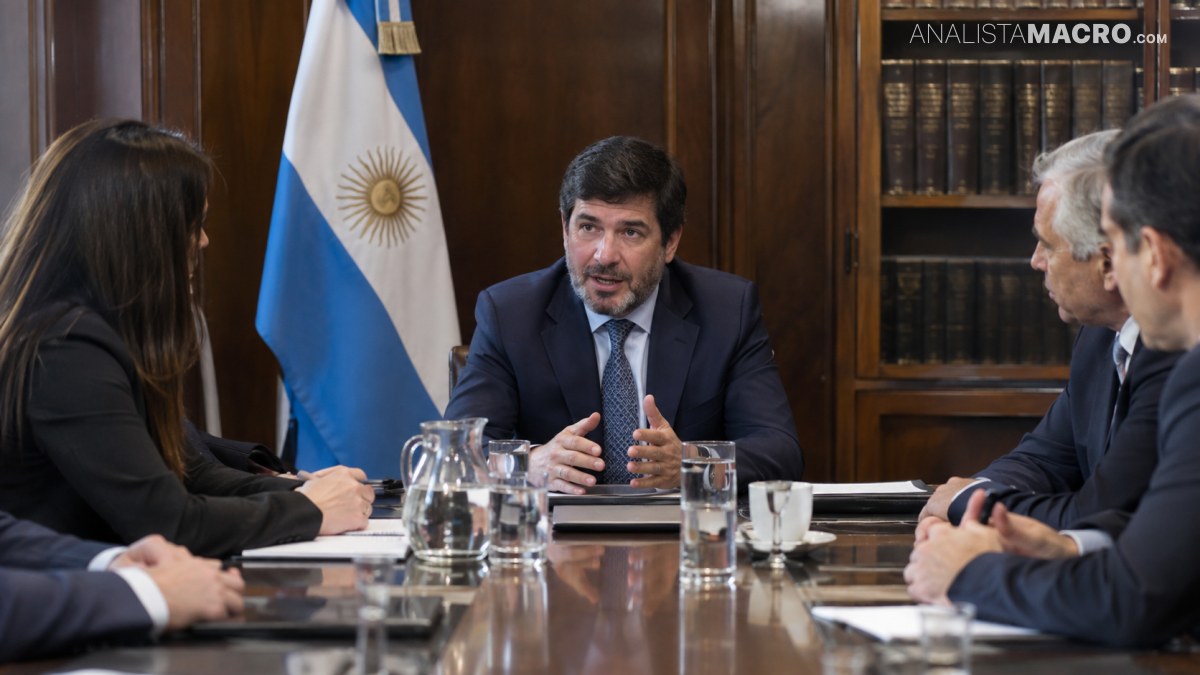 Cinco personas se reúnen alrededor de una mesa, con un hombre hablando frente a una bandera argentina y estanterías de libros.