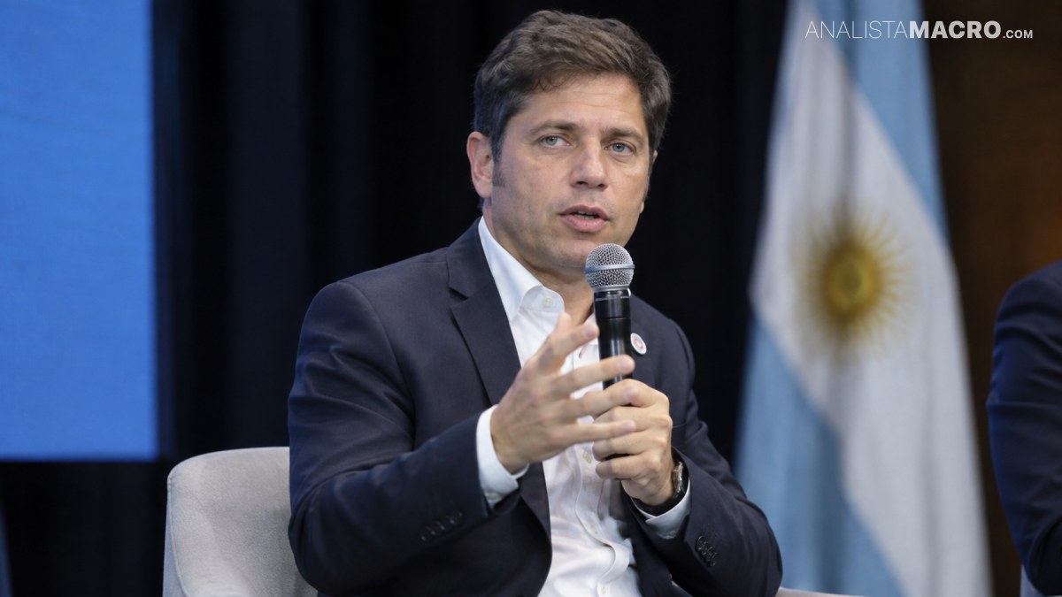 Hombre con traje oscuro sostiene un micrófono y gesticula, sentado frente a una bandera argentina y cortinas negras.