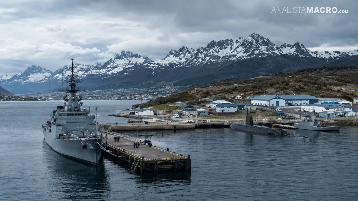 Barco militar atracado en un puerto junto a casas costeras, con montañas nevadas y nubes en el fondo.