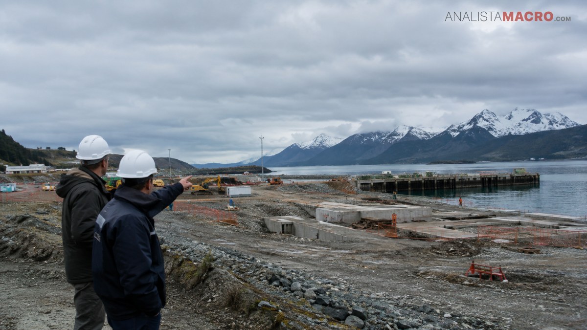 Dos personas con cascos observan un muelle industrial junto a un lago, rodeado de montañas nevadas bajo un cielo nublado.