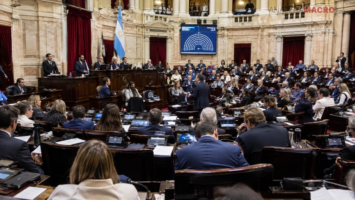 Numerosas personas ocupan los escaños de una sala parlamentaria, frente a una mesa presidencial y una pantalla iluminada.