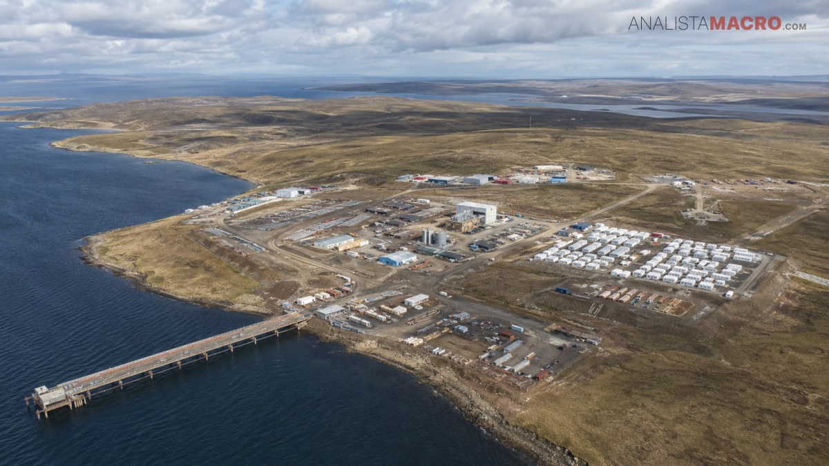 Vista aérea de un complejo industrial junto a una costa, con muelle, edificios, depósitos y extensos terrenos áridos bajo nubes.