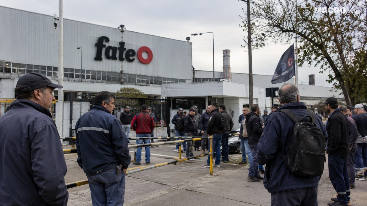 Personas esperan frente a un edificio industrial con el logotipo “fate”, mientras varias llevan banderas y mochilas.