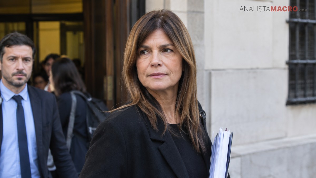 Mujer de cabello castaño sostiene varios documentos frente a un edificio, acompañada por otras personas.