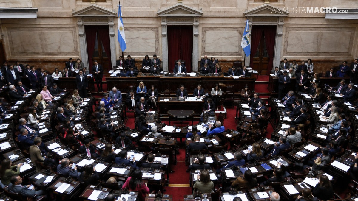 Salón legislativo amplio con numerosos funcionarios sentados en escritorios, banderas y una mesa central rodeada de asientos.