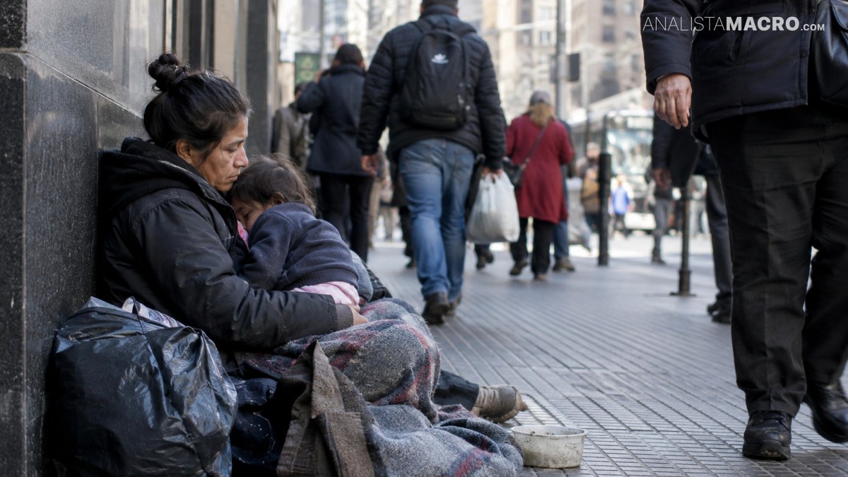 Varias personas caminan por una calle; una mujer y un niño están sentados junto a una pared, junto a una bolsa.