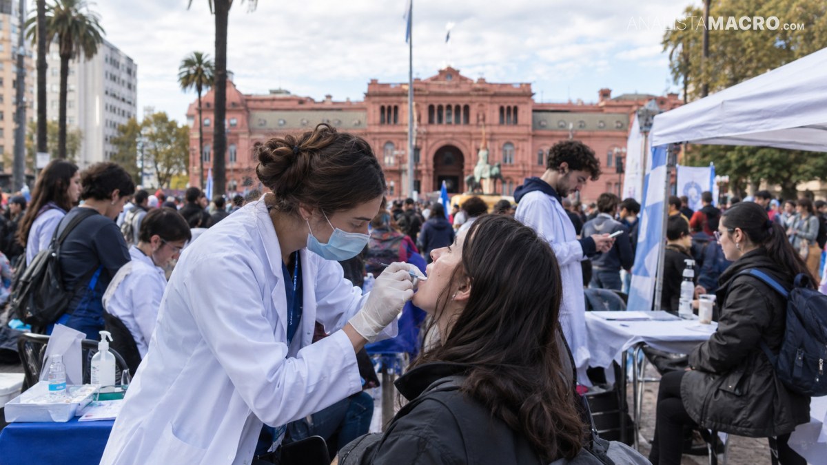 Una profesional sanitaria examina la boca de una mujer mientras varias personas esperan en puestos al aire libre frente a un edificio rosado.
