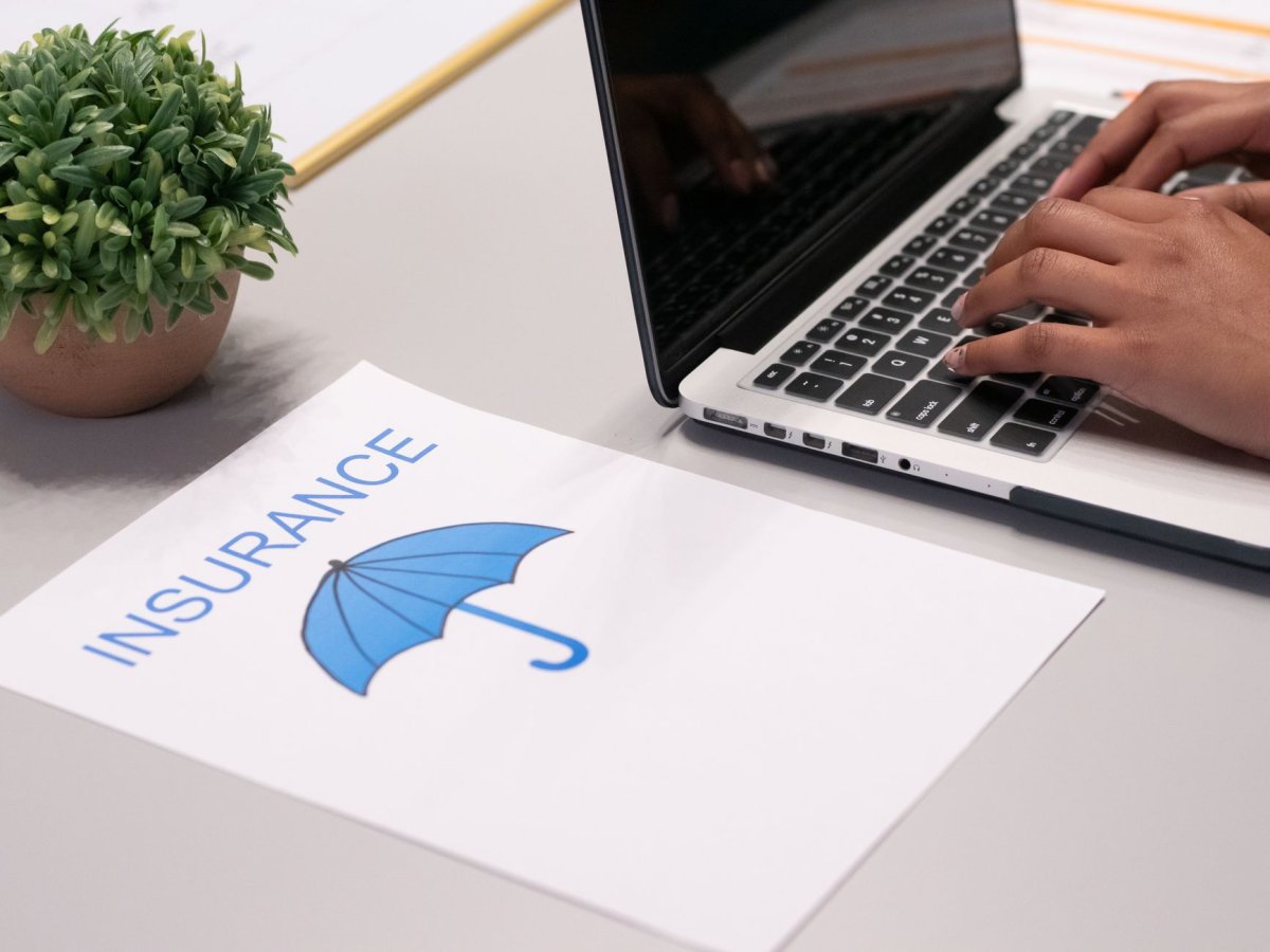 Una mano escribe en un portátil sobre un papel con el texto “INSURANCE” y un icono de paraguas.