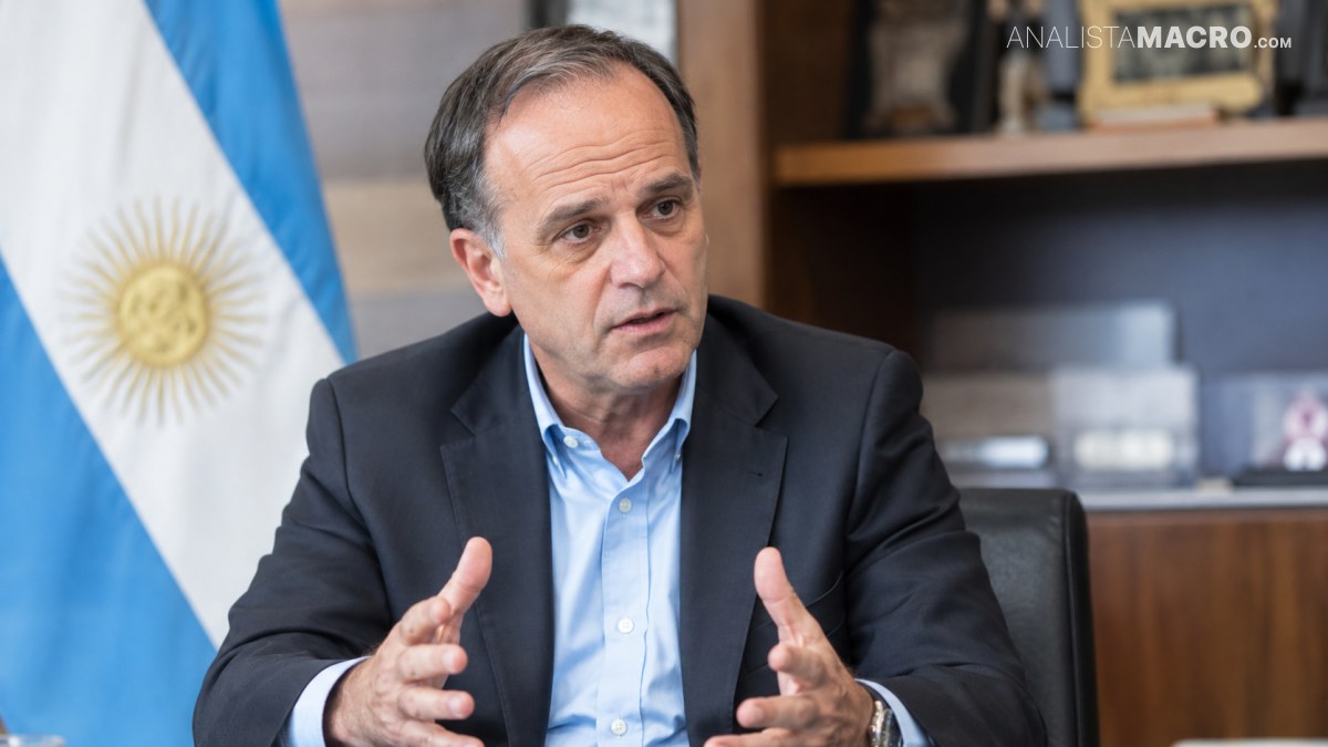 Hombre de cabello oscuro con saco y camisa azul, sentado y gesticulando frente a una bandera argentina.
