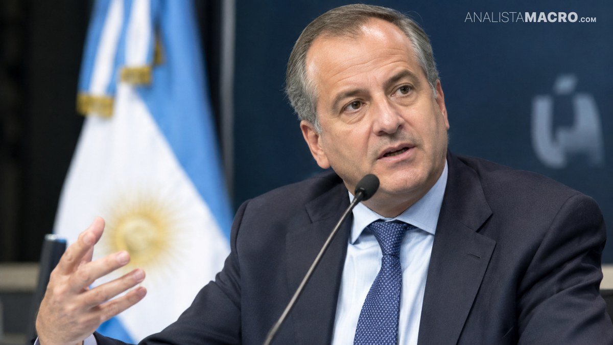 Hombre de traje y corbata habla ante un micrófono, gesticulando con una mano frente a una bandera argentina.