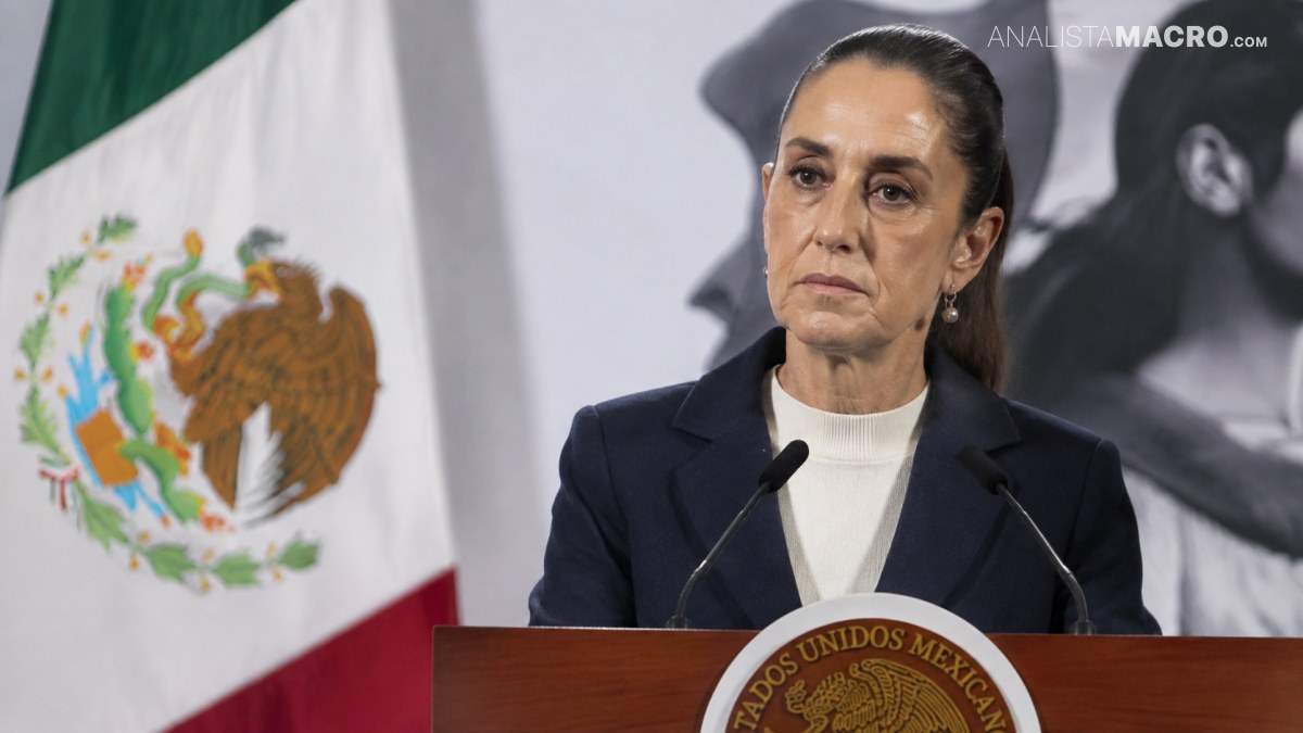 Mujer con traje oscuro y cuello blanco habla ante un atril, con banderas y una imagen en blanco y negro al fondo.