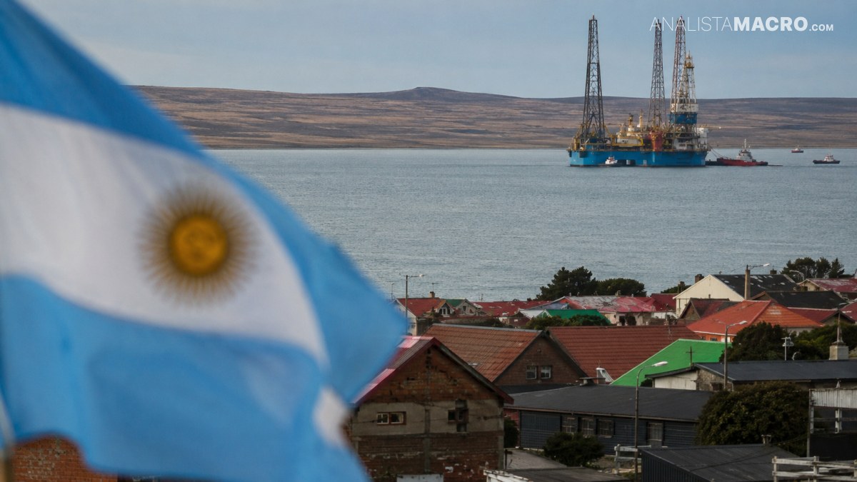 Bandera argentina desenfocada en primer plano, viviendas costeras y plataformas de perforación mar adentro bajo un cielo despejado.