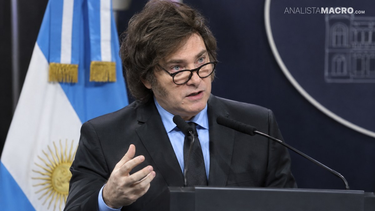 Hombre con gafas y traje oscuro habla ante un micrófono, gesticulando frente a banderas y un fondo institucional.