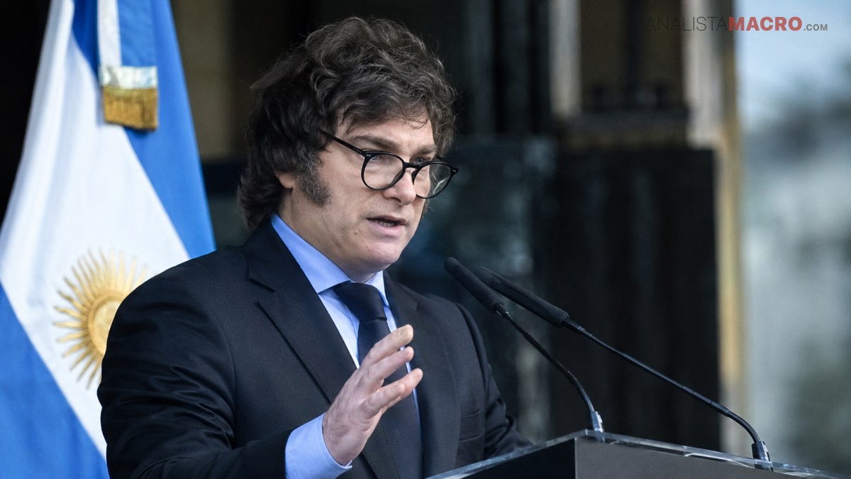 Un hombre con gafas y traje habla ante dos micrófonos, con una bandera argentina visible detrás.