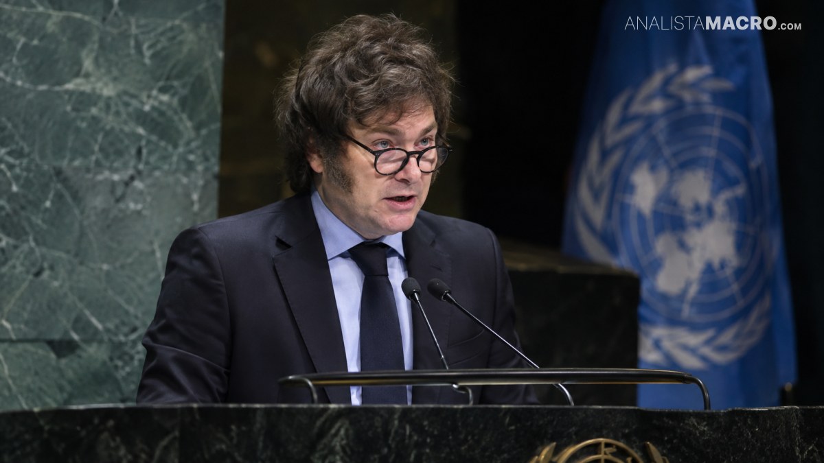 Persona con gafas habla en un podio con micrófonos, con bandera azul y pared verde al fondo.