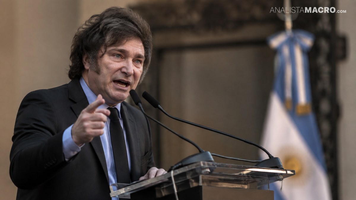Hombre de cabello oscuro habla ante micrófonos, gesticulando junto a un atril, con una bandera celeste y blanca al fondo.