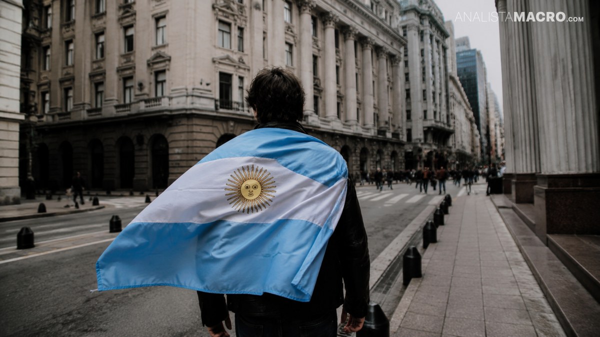 Persona envuelta en una bandera argentina camina por una calle flanqueada por imponentes edificios urbanos.