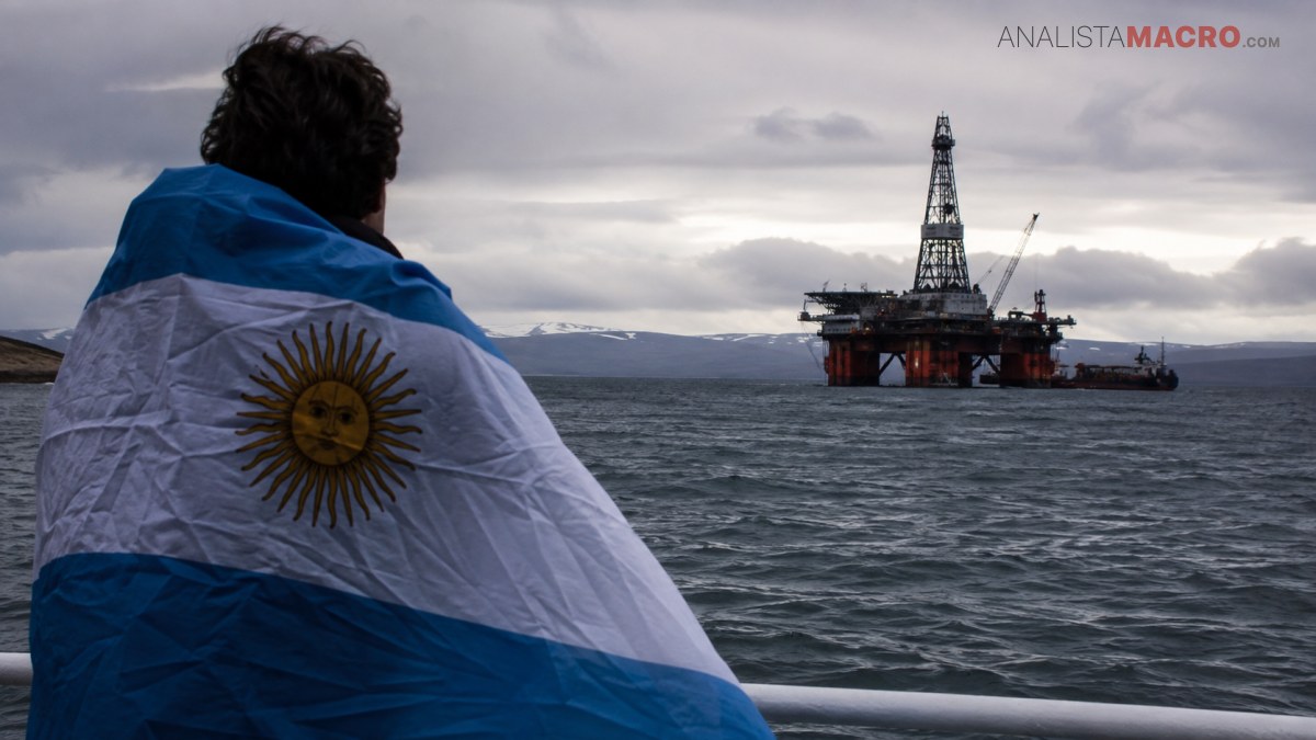 Persona envuelta en una bandera argentina observa una plataforma petrolera sobre el mar bajo un cielo nublado.