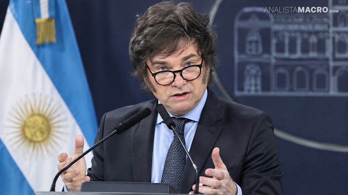 Hombre con gafas y traje oscuro habla ante un micrófono, con una bandera celeste y blanca al fondo.
