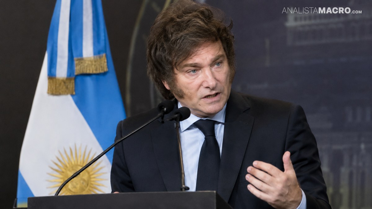 Un hombre con traje oscuro habla ante un micrófono, con una bandera argentina visible detrás y una mano levantada.
