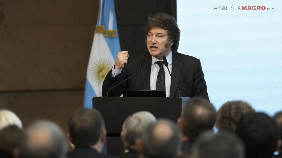 Hombre de cabello oscuro habla ante un micrófono desde un atril, con una bandera celeste y blanca detrás.
