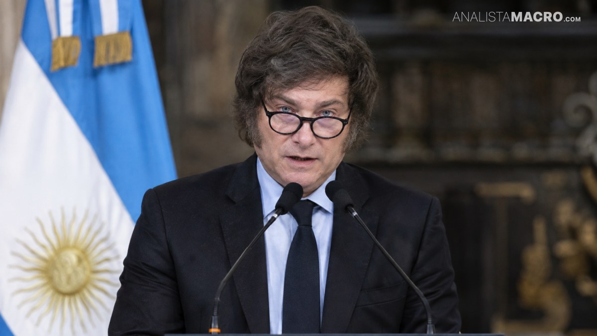 Hombre con gafas y traje oscuro habla ante dos micrófonos, con una bandera celeste y blanca detrás.