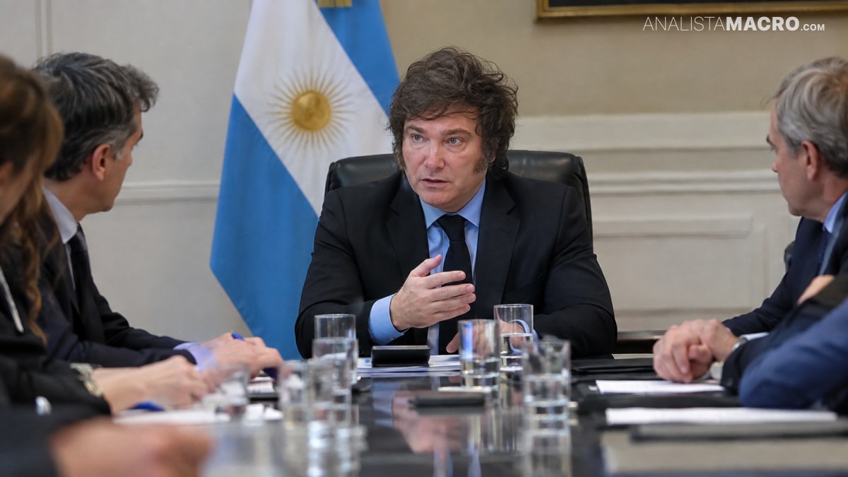 Hombre sentado al centro gesticula durante una reunión, acompañado por varias personas y una bandera argentina al fondo.