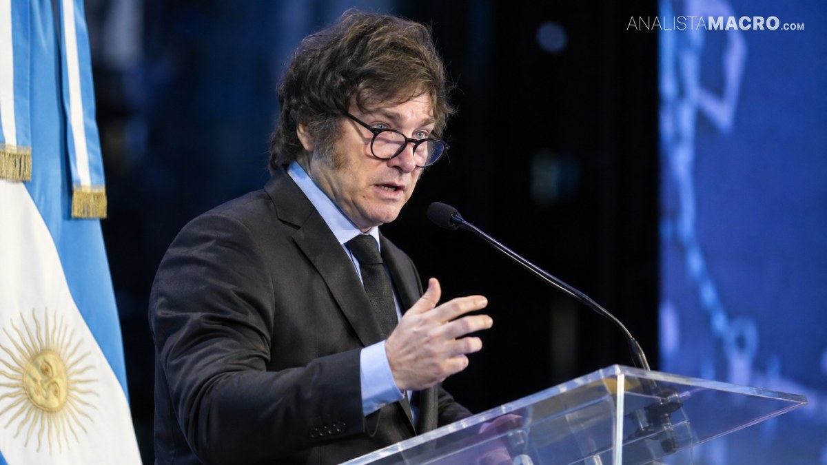 Hombre con traje oscuro y gafas habla en un atril con micrófono, junto a banderas y fondo azul.