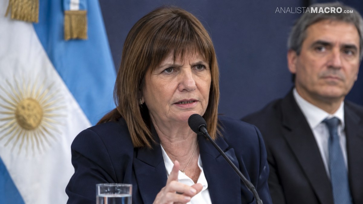 Mujer de cabello castaño habla ante un micrófono y señala con un dedo, mientras un hombre y una bandera aparecen detrás.