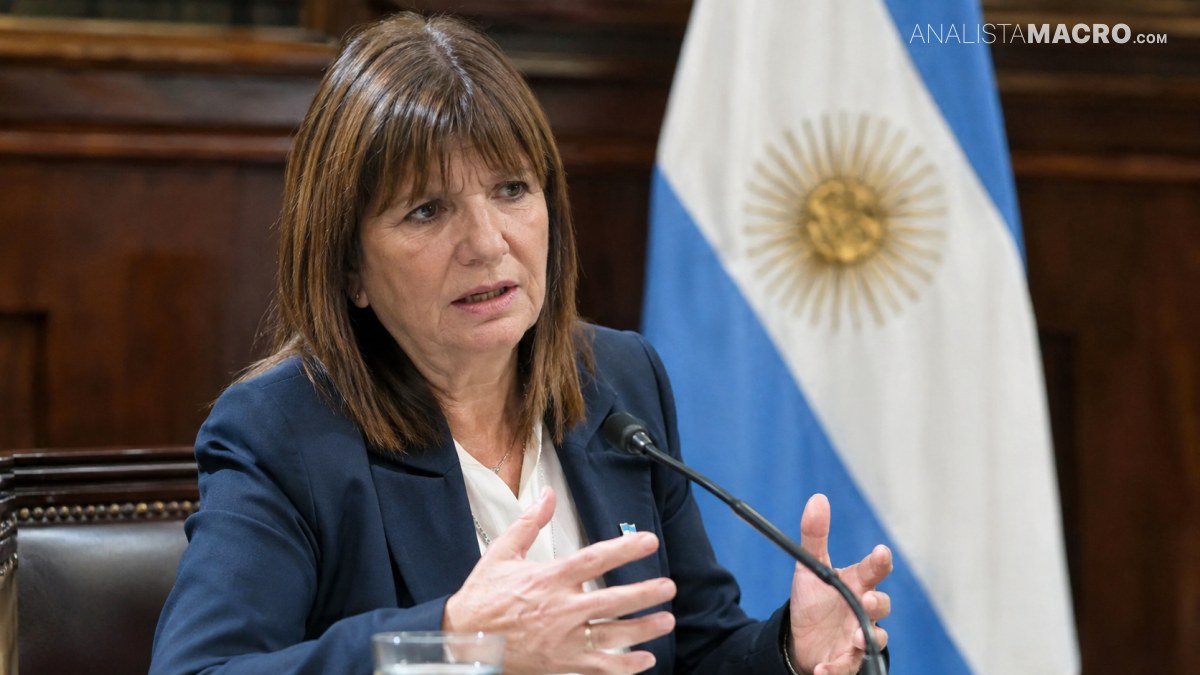 Mujer de cabello castaño, vestida con saco azul, habla y gesticula junto a un micrófono frente a banderas.