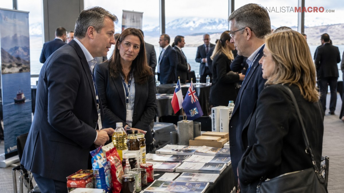 Cuatro personas conversan junto a una mesa con folletos, productos, botellas y dos pequeñas banderas, mientras se observan montañas al fondo.