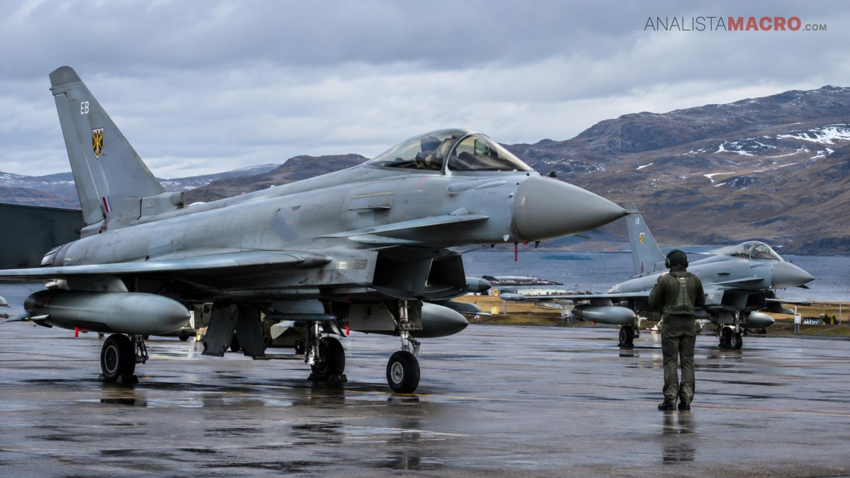 Dos aviones de combate estacionados en una pista mojada, con montañas nevadas y una persona caminando al fondo.